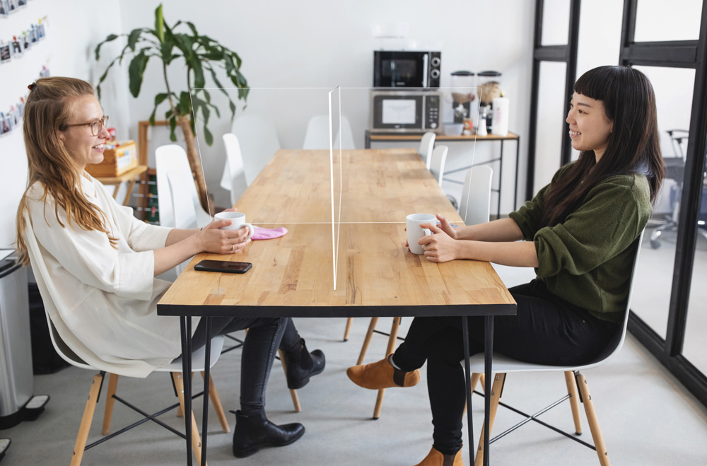 A clear transparent 4-way divider between two women smiling, sipping coffee at a table.