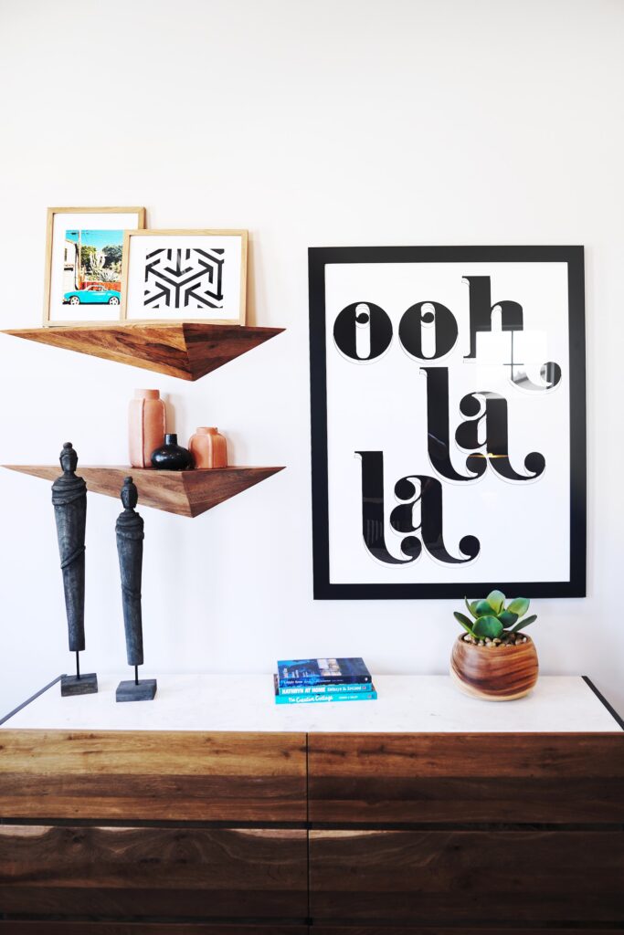Wood dresser with statues, books, and a plant below a "ooh la la" black and white sign and floating wooden shelves.