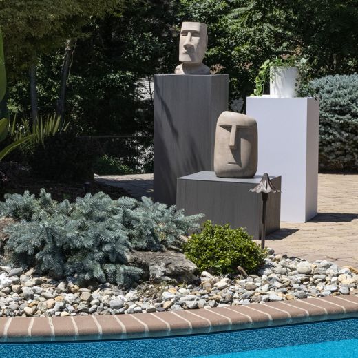 Three outdoor display pedestals holding statues and a plant in different heights behind a pool
