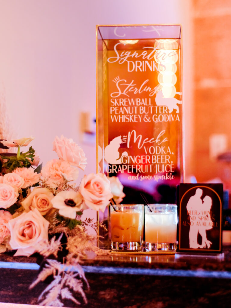 Drinks table at a wedding with an acrylic sign featuring signature drinks and ingredients.