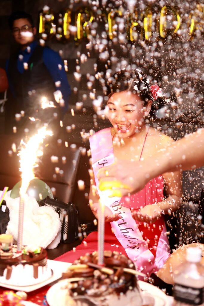 Woman sitting in a birthday sash smiling at birthday cake with a big and bright candle.